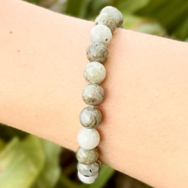 A close-up of a wrist wearing the Spellcrafter Labradorite Crystal Bracelet, featuring round, polished grey-green stones against a blurred green background.