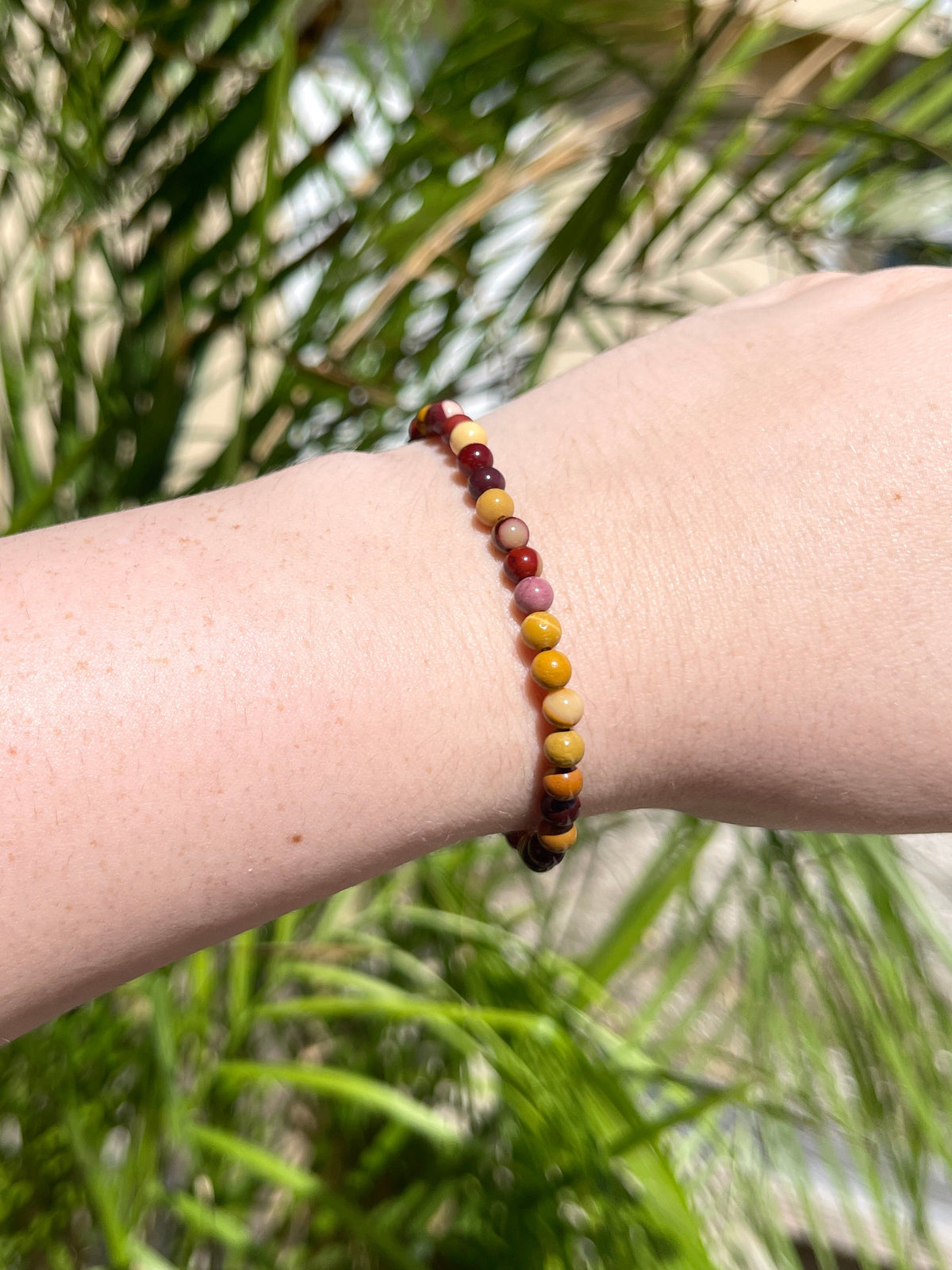 A close-up of a wrist wearing the Spellcrafter Mookaite Jasper Bracelet, featuring thin crystal beads in red, yellow, and brown. Blurred green foliage in bright sunlight highlights the healing energy of these vibrant stones.