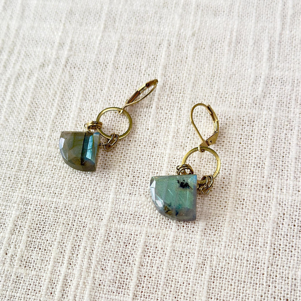 The Spellcrafter Labradorite Diamond Earrings feature hypoallergenic brass lever-backs and geometric blue-green gemstone pendants, beautifully displayed on textured cream fabric.