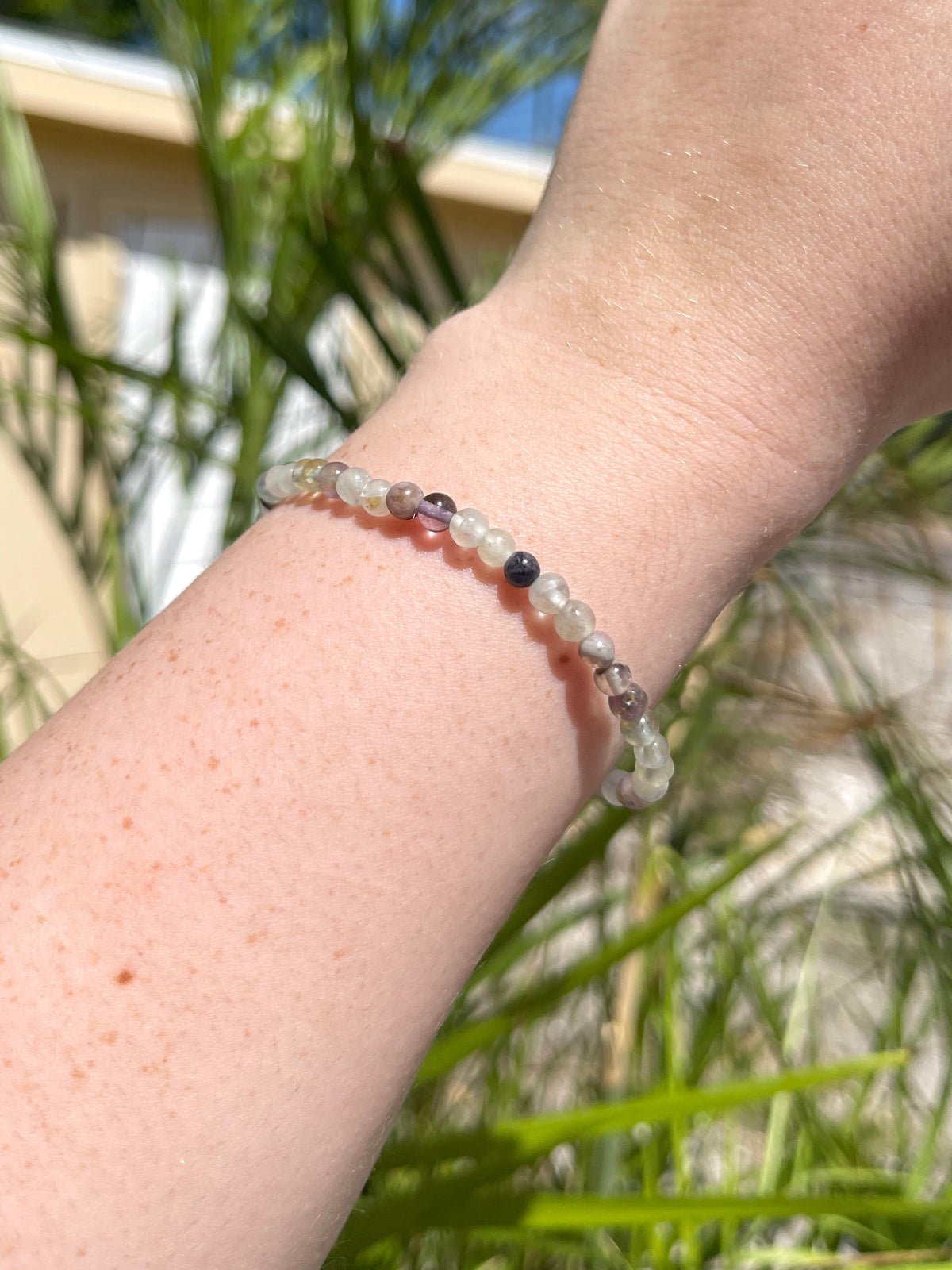 A close-up of a wrist adorned with the Spellcrafter Fluorite Crystal Bracelet, featuring small multicolored stones. Green plants and the sunlit exterior of a beige building are visible in the background.