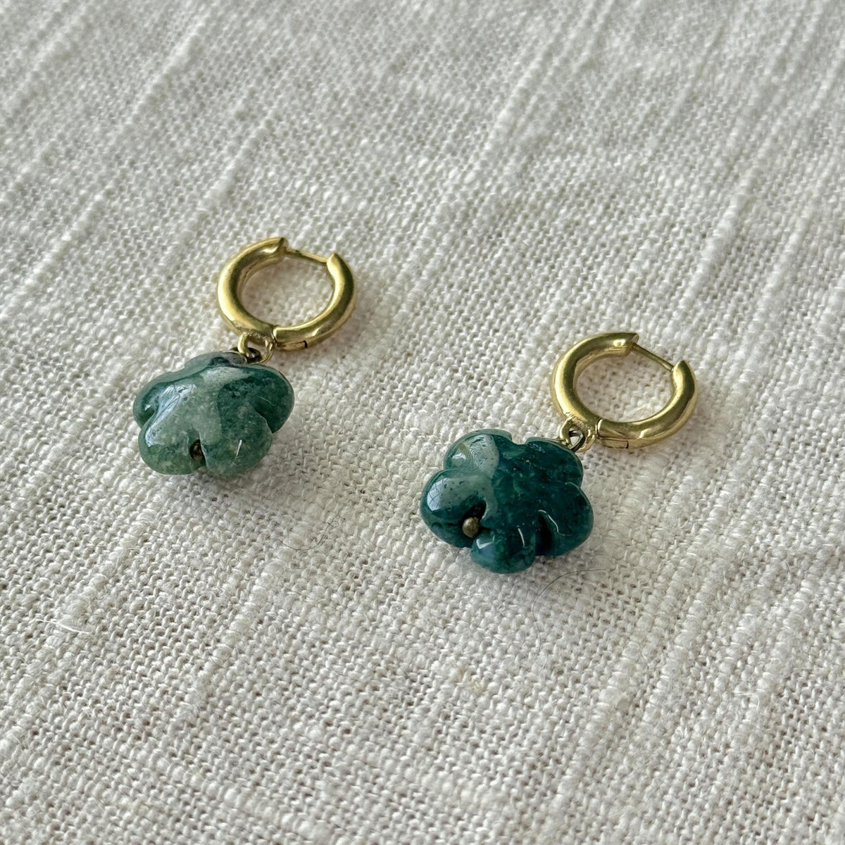 The Spellcrafter Moss Agate Flower Earrings, gold hoop and hypoallergenic, with green four-leaf clover pendants, rest on textured off-white fabric.