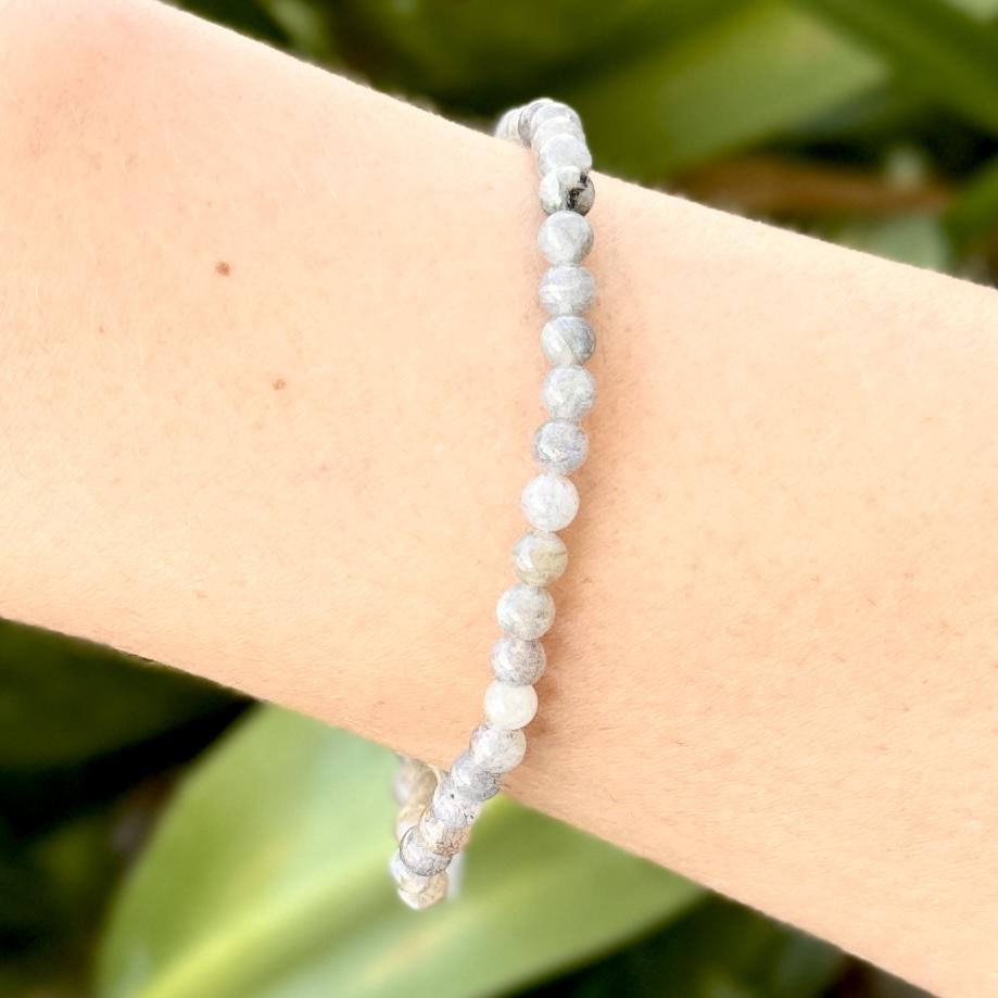 A close-up of a wrist adorned with Spellcrafter’s Labradorite Crystal Bracelet, featuring small, round, light gray beads. The blurred green background accentuates this elegant piece renowned for its crystal healing properties.