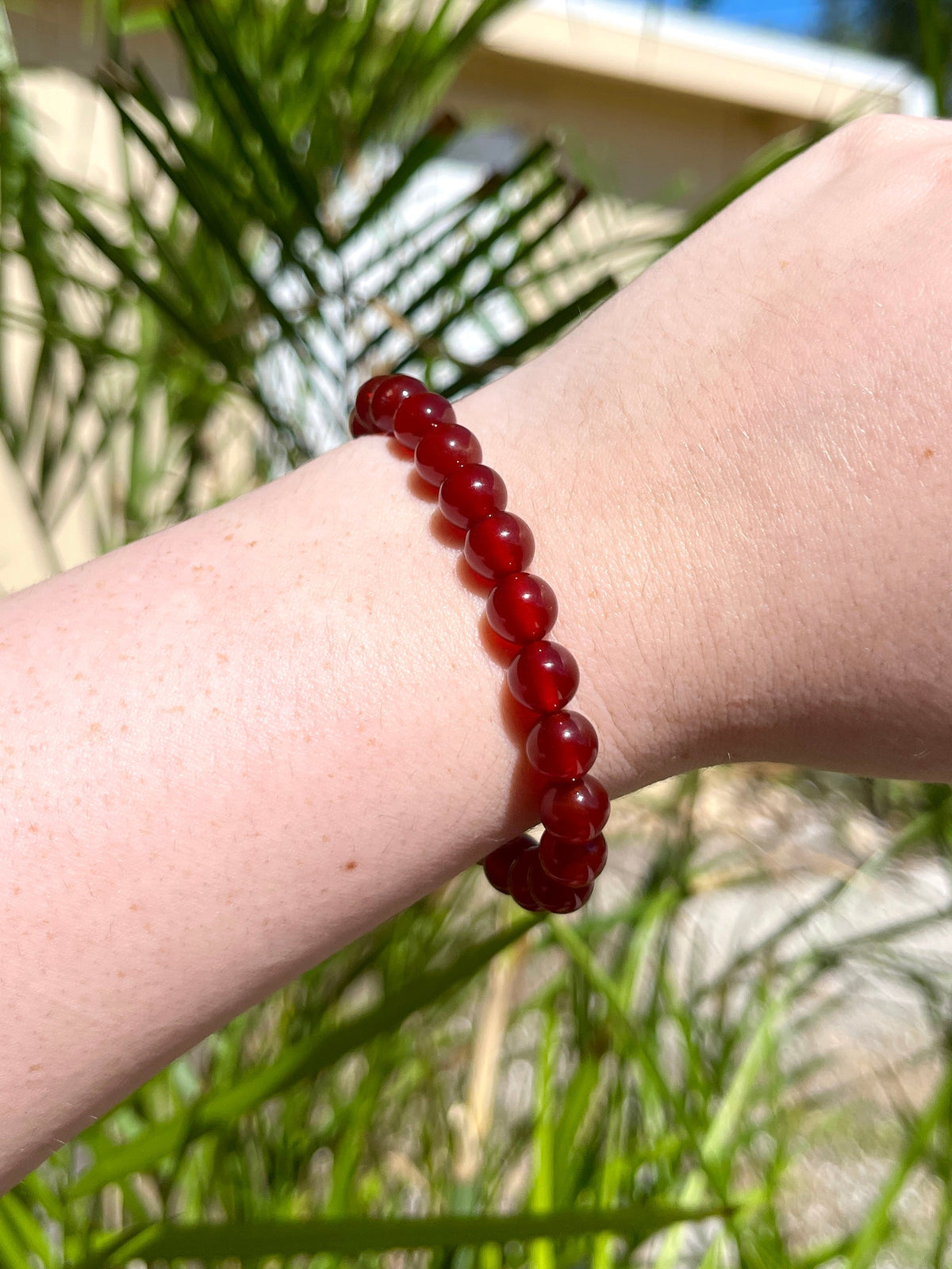 A fair-skinned hand wears the Spellcrafter Carnelian Crystal Bracelet, featuring round, glossy red beads, showcased outdoors with green plants and a light building in the background.