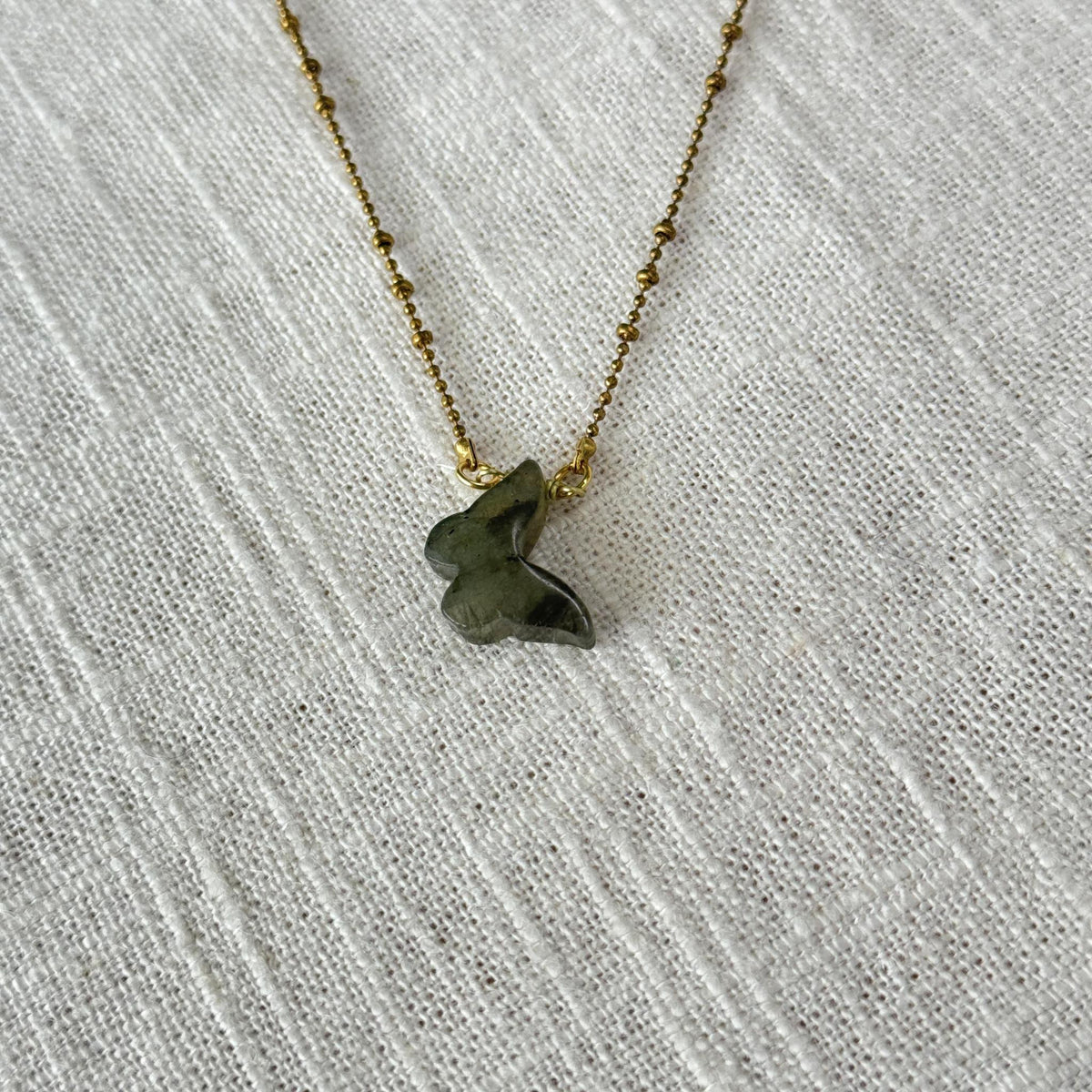 The Spellcrafter Labradorite Butterfly Necklace showcases a delicate gold chain with a small green labradorite butterfly pendant, displayed on textured white fabric.