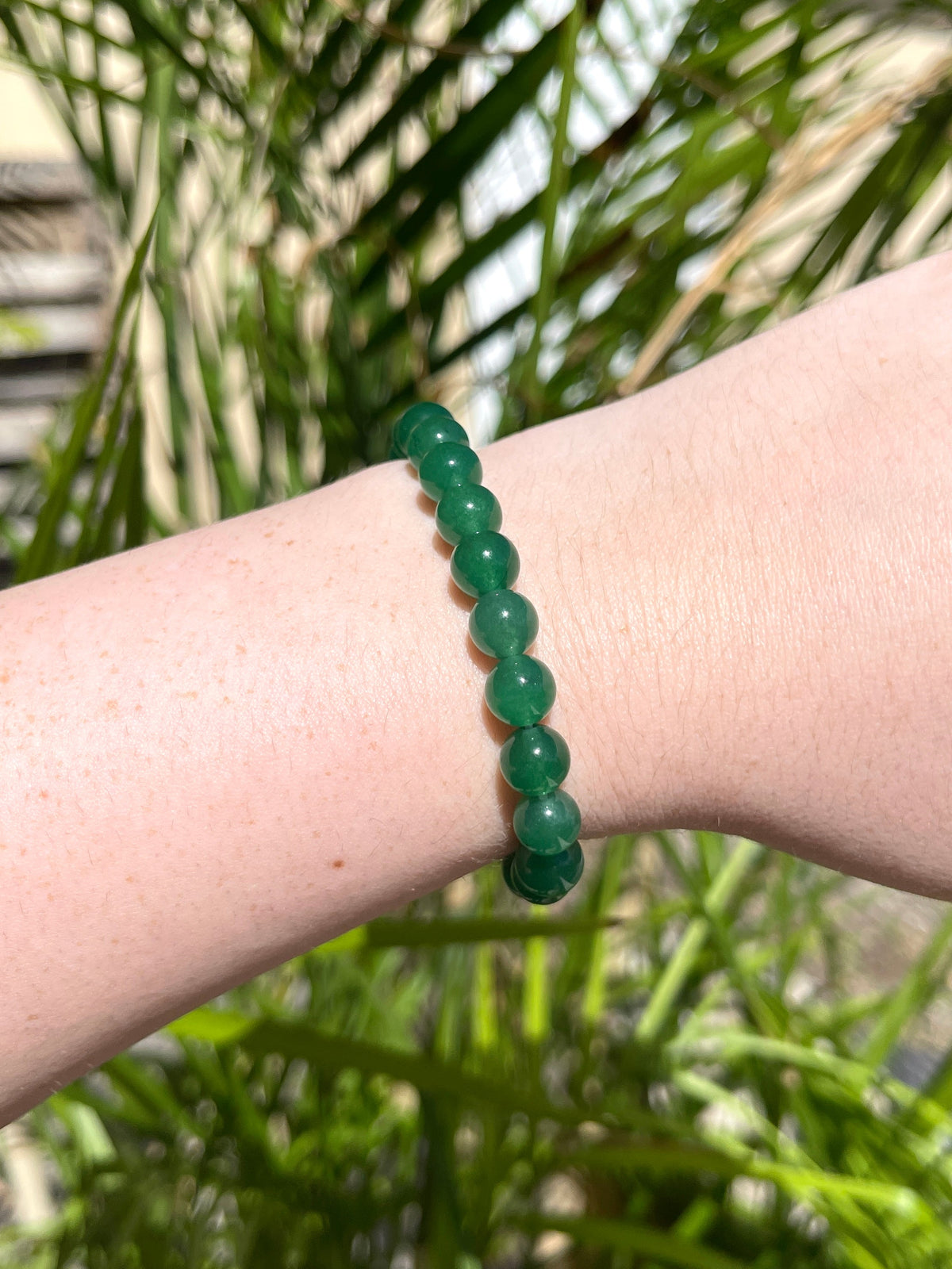 A wrist adorned with the Spellcrafter Green Aventurine Bracelet—featuring round green beads—gleams under bright sunlight, with palm leaves in the background, evoking a sense of healing energy.