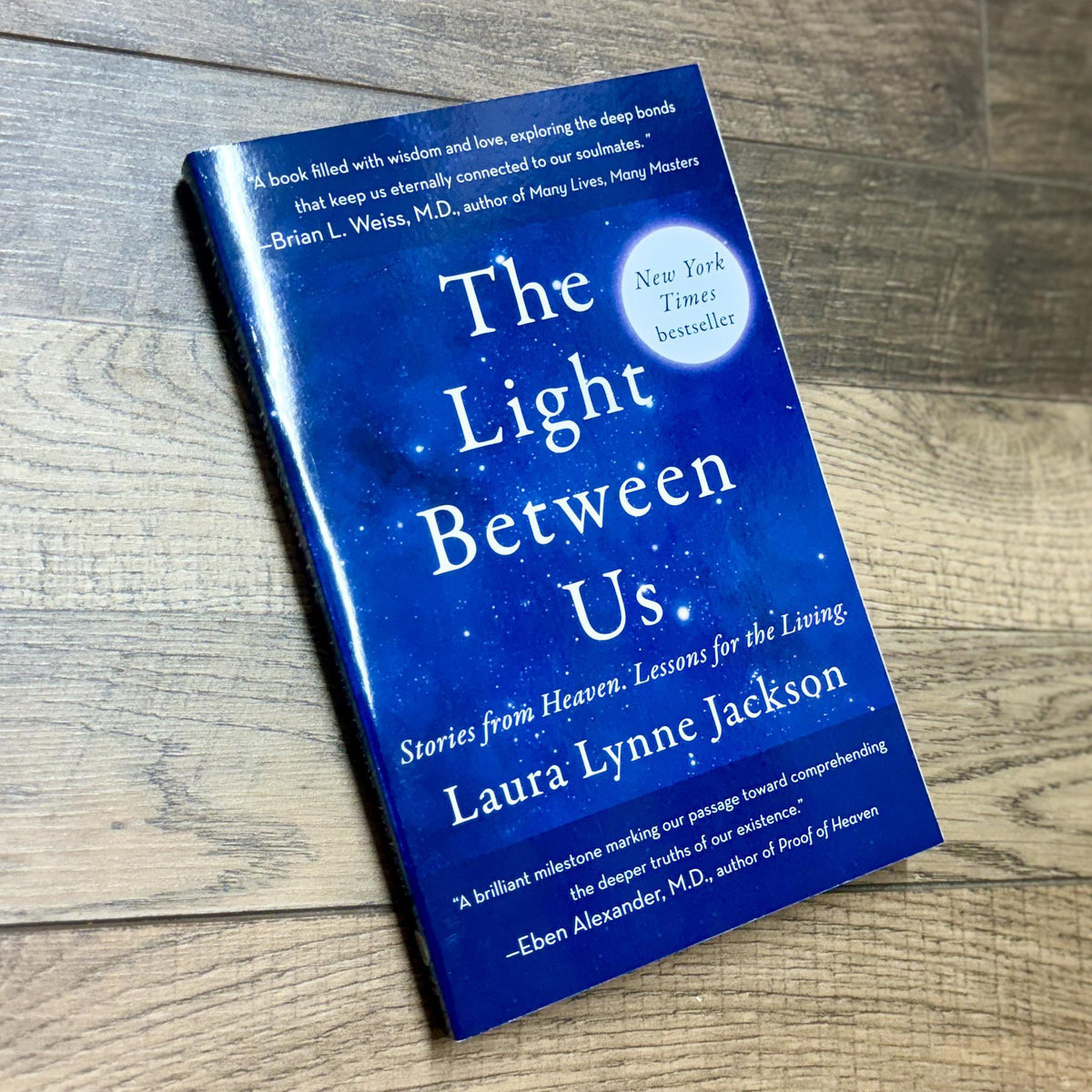 The Light Between Us by Spellcrafter rests on a wooden floor, its blue cover adorned with white and yellow text, promotional blurbs, and a New York Times bestseller badge.