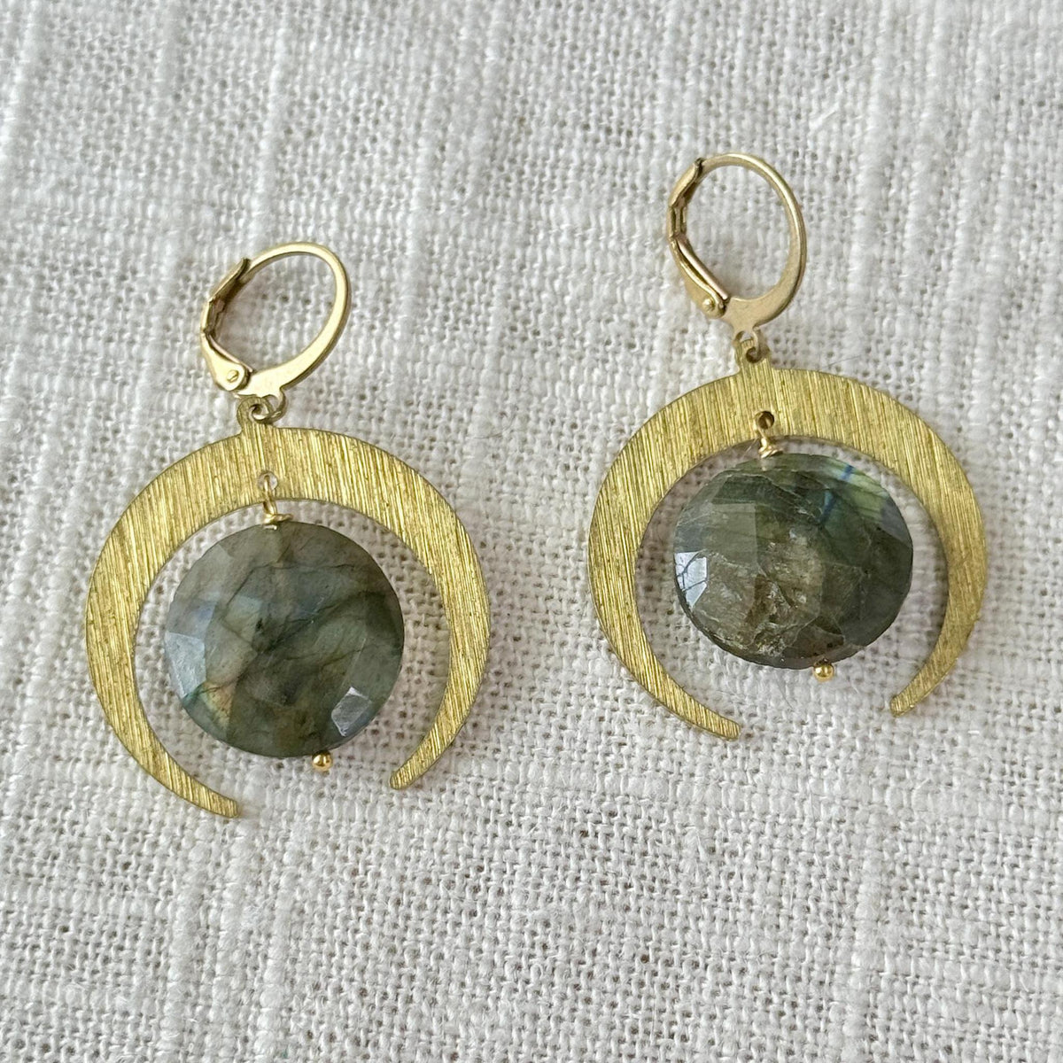 The Spellcrafter Labradorite Lunar Eclipse Earrings feature gold crescent moon dangles with a textured finish and central round grey-green labradorite gems, elegantly displayed on light woven fabric.