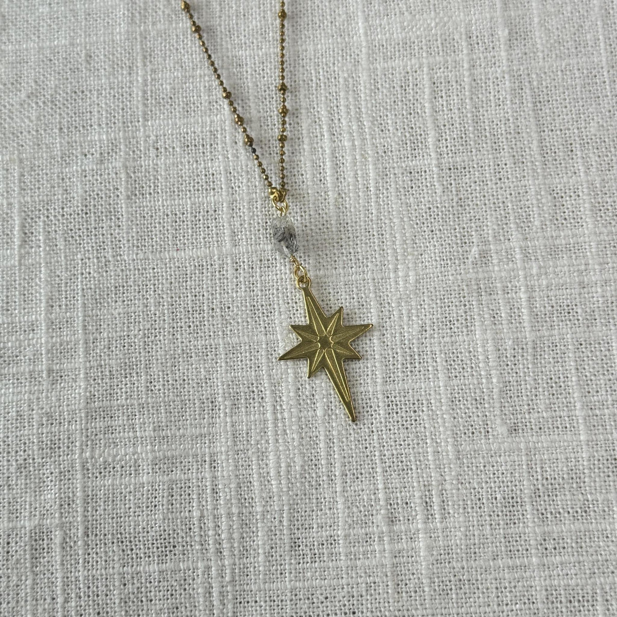 The Spellcrafter Herkimer Diamond North Star Necklace, featuring a star-shaped pendant and a small Herkimer Diamond bead, is displayed on a textured white fabric background.