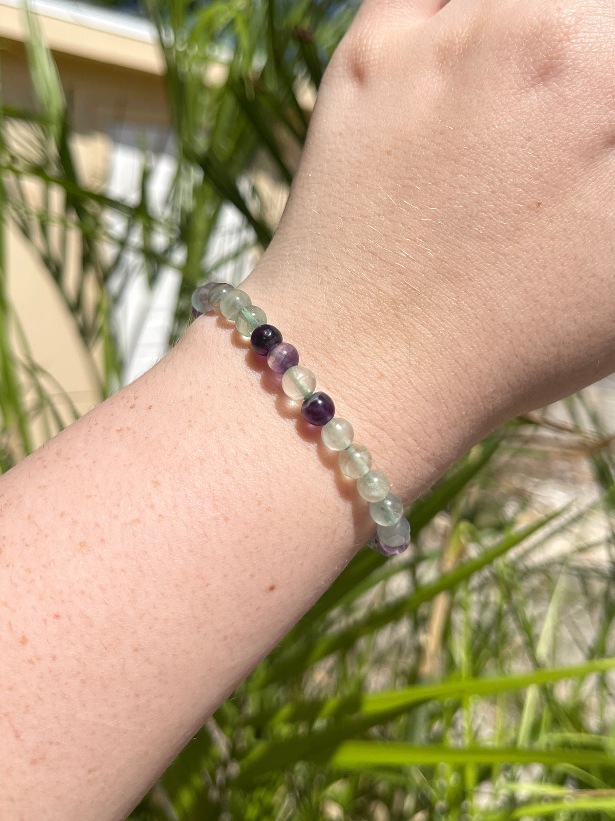 A close-up of a hand wearing the Spellcrafter Fluorite Crystal Bracelet, with translucent green and purple beads, outdoors in bright sunlight, green plants and a beige building blurred in the background.