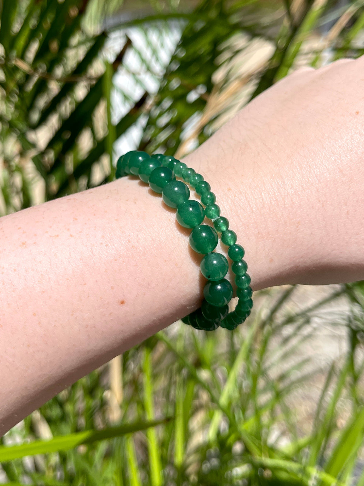 A fair-skinned hand wears two green beaded bracelets, including the Spellcrafter Green Aventurine Bracelet, shown outdoors with blurred palm leaves in the background, radiating healing energy.