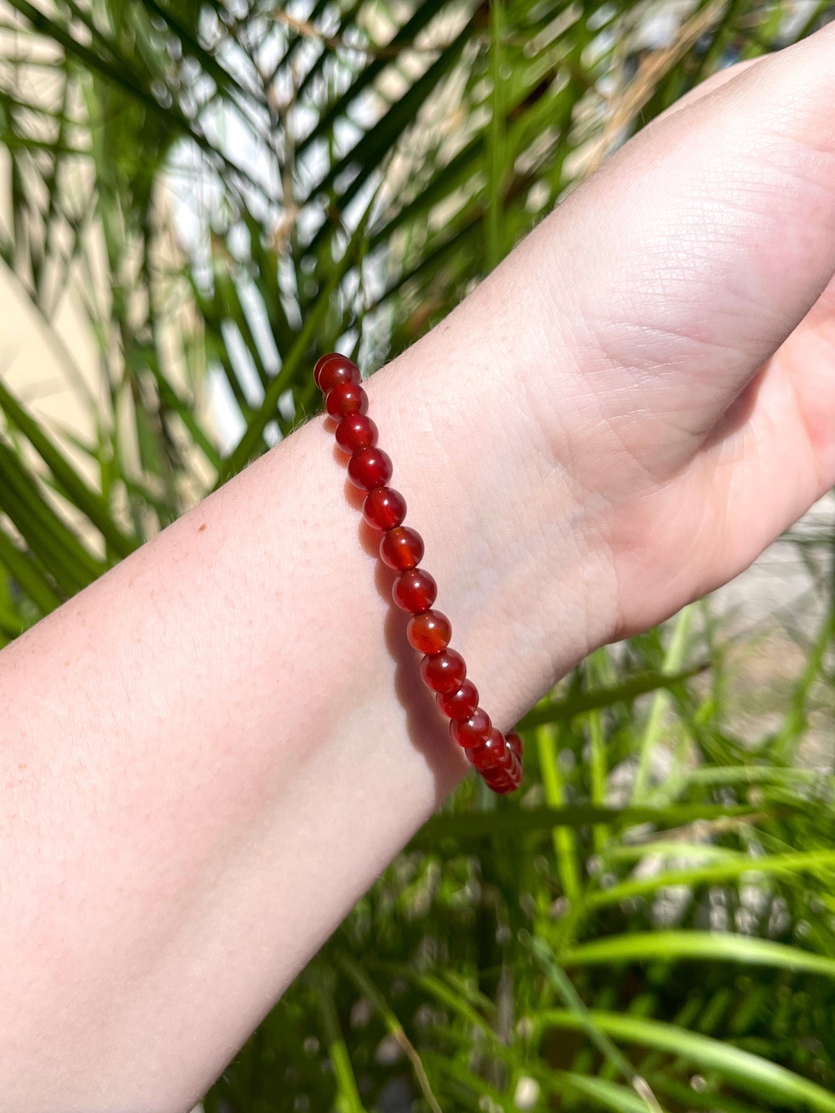 A hand wearing the Spellcrafter Carnelian Crystal Bracelet, made of small round red beads, is shown in bright sunlight against a backdrop of green leaves and blurred outdoor scenery.
