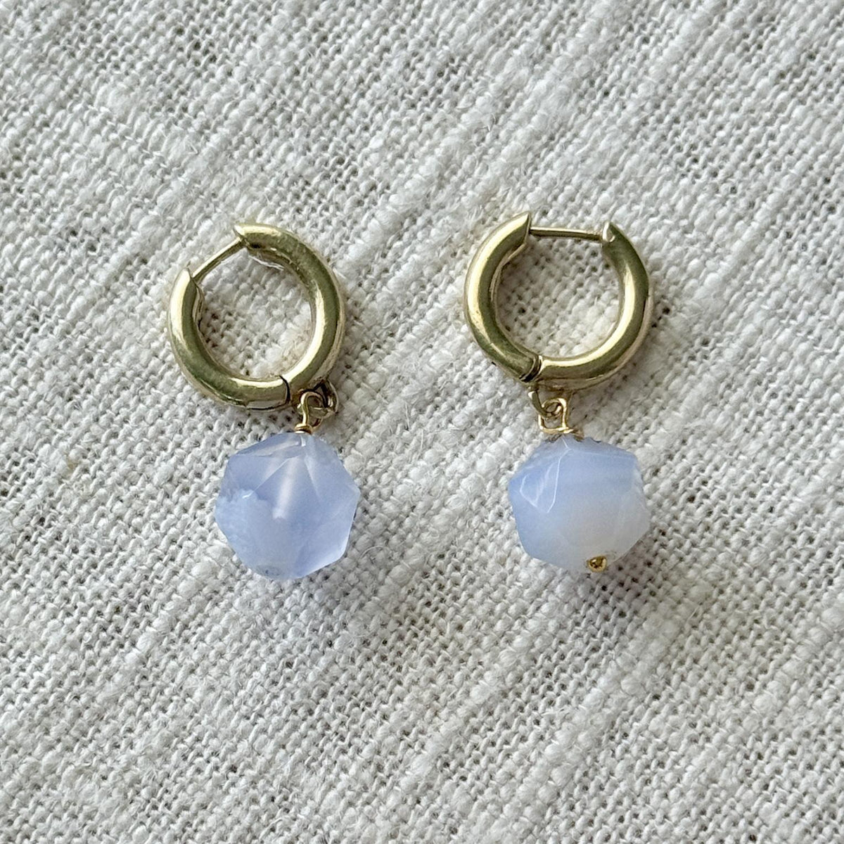 The Spellcrafter Blue Lace Agate Gold Huggie Hoop Earrings feature faceted blue gemstone beads on small gold hoops, shown here against a textured off-white fabric background.