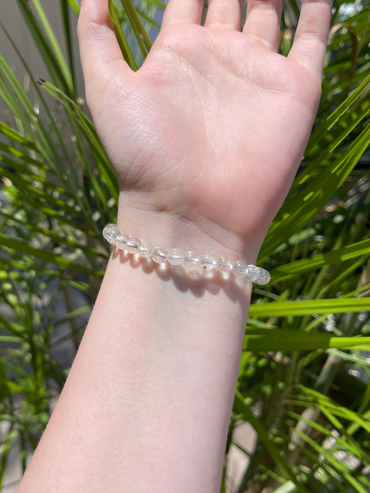 A hand wearing the Spellcrafter Cracked Clear Quartz Bracelet is raised in sunlight, with softly blurred green spiky plant leaves in the background.