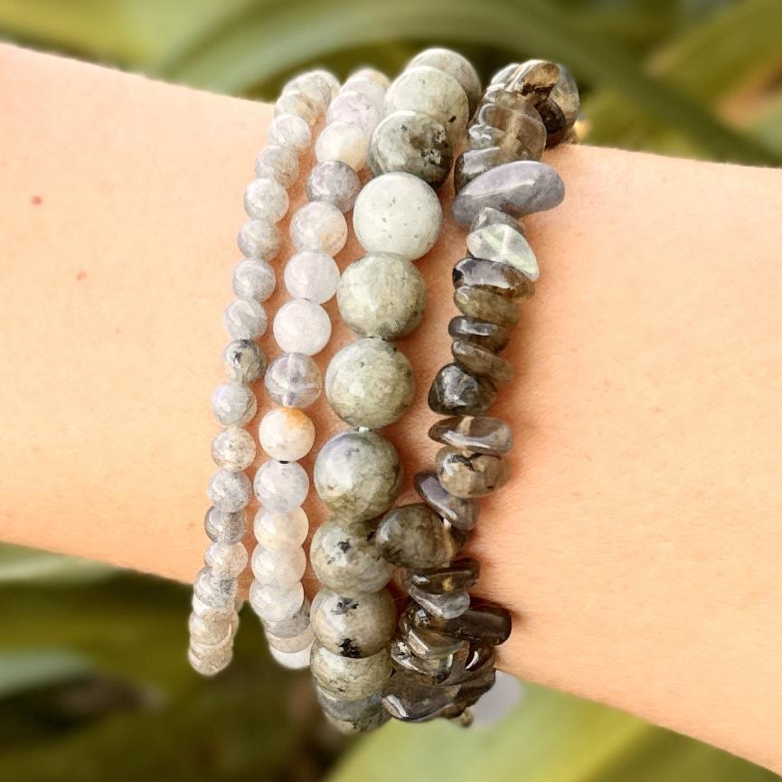 A close-up of a forearm wearing the Spellcrafter Labradorite Crystal Bracelet, featuring round and irregular natural stone beads in gray, green, and brown tones, with a soft-focus green background.