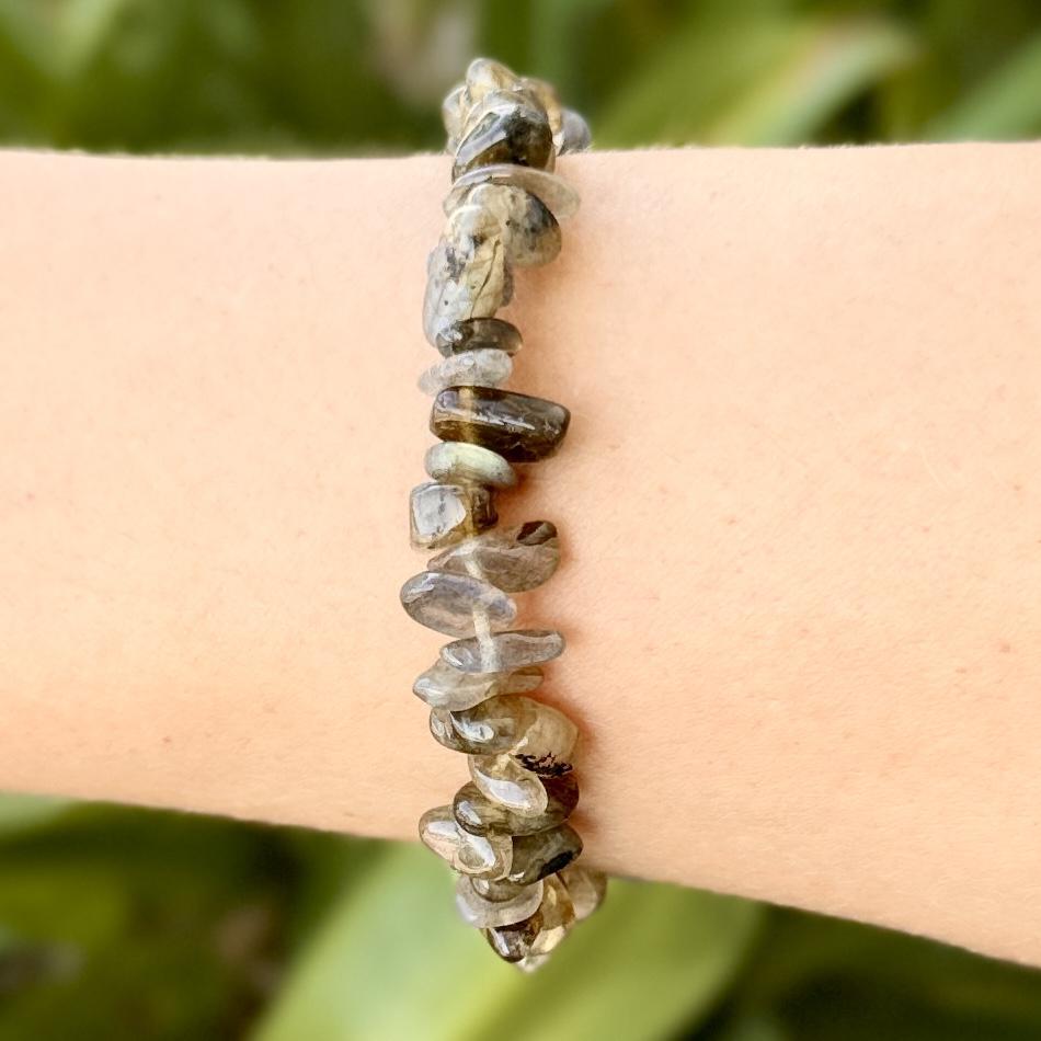 A close-up of a wrist adorned with the Spellcrafter Labradorite Crystal Bracelet, featuring polished, irregular brown and gray stones, set against a blurred green background.