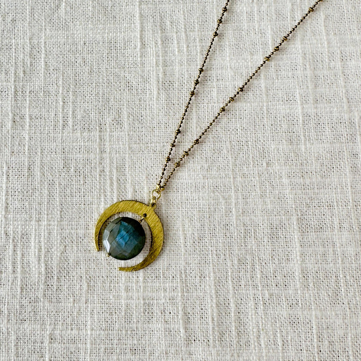 The Spellcrafter Labradorite Lunar Eclipse Necklace showcases a crescent moon pendant with a faceted dark blue-green labradorite, elegantly displayed on textured off-white fabric.