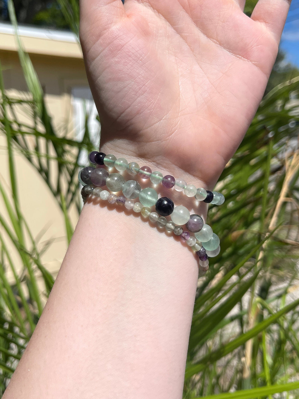 A hand with light skin is shown outdoors wearing four Spellcrafter Fluorite Crystal Bracelets, featuring translucent green and purple stones, with green plants and a beige building in the background.