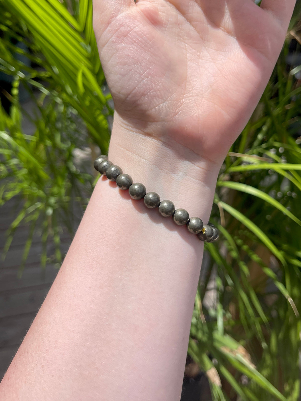A hand wearing the Spellcrafter Pyrite Crystal Bracelet is raised in sunlight, with green leafy plants softly blurred in the background.