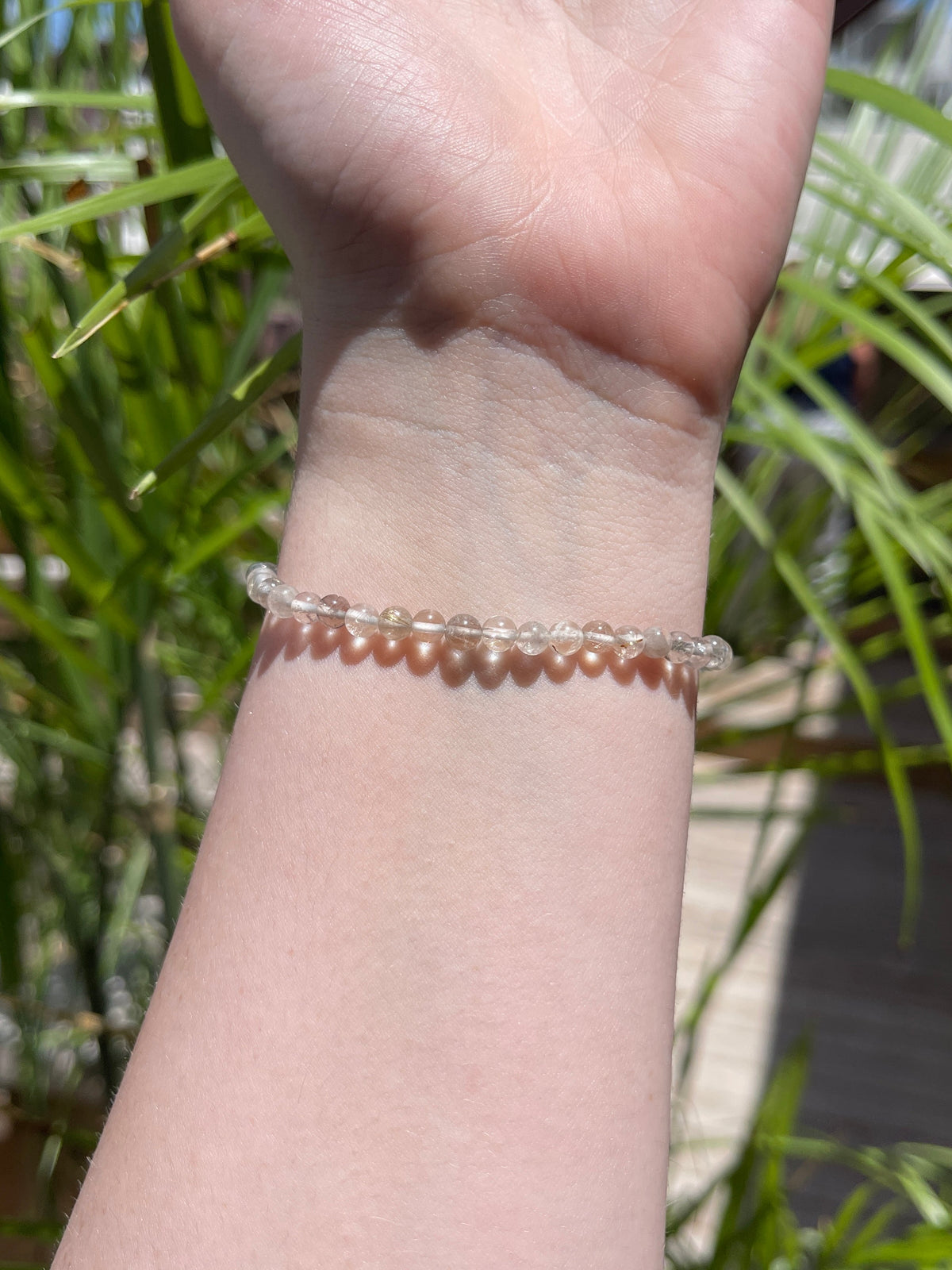 A close-up of a wrist adorned with the Spellcrafter Cracked Clear Quartz Bracelet, sunlight casting bead-shaped shadows on the skin. Blurred green leaves in the background highlight its healing energy.
