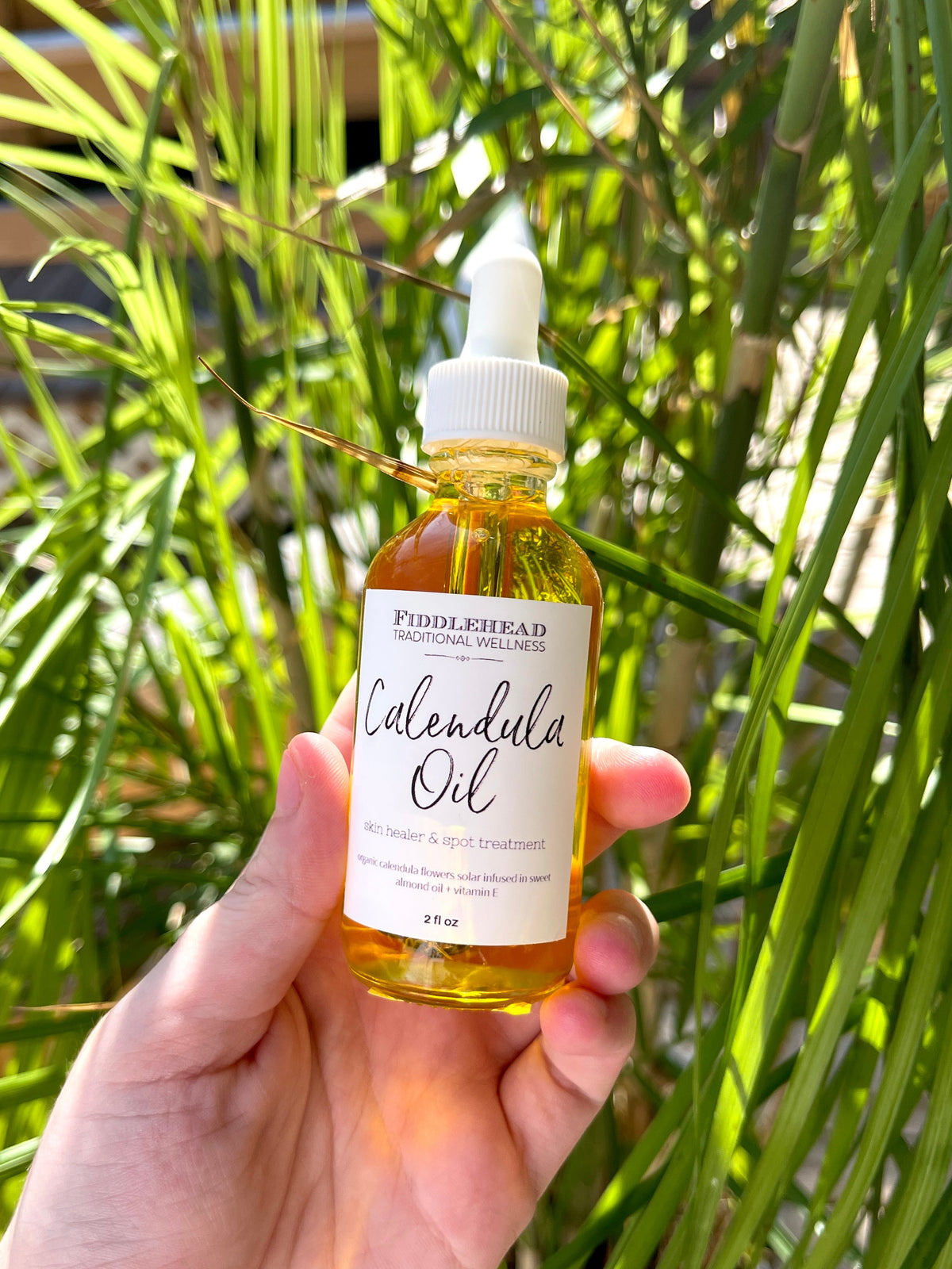 A hand holds a dropper bottle labeled Calendula Oil by Spellcrafter—this herbal infused oil glows in sunlight, set against a backdrop of vibrant green plants.