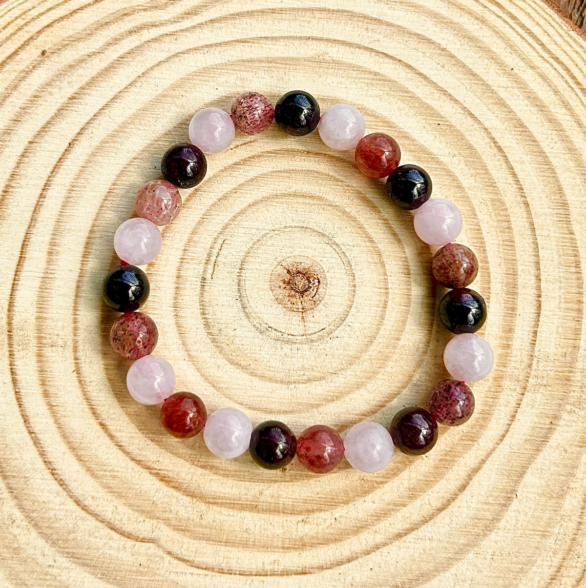The Spellcrafter Heart Chakra Trio Crystal Bracelet, with alternating pink and dark red stones, is displayed in a circle on wood with visible tree rings—ideal for balancing your heart chakra.