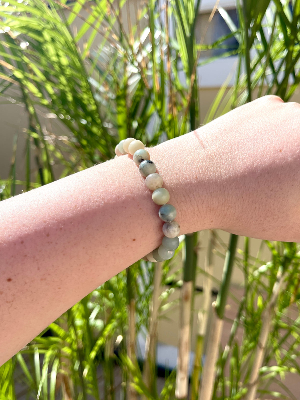 A light-skinned arm wears the Spellcrafter Sunstone Crystal Bracelet—stretchy with green, white, and gray stones—held up outdoors among tall plants and a blurred building in the background.