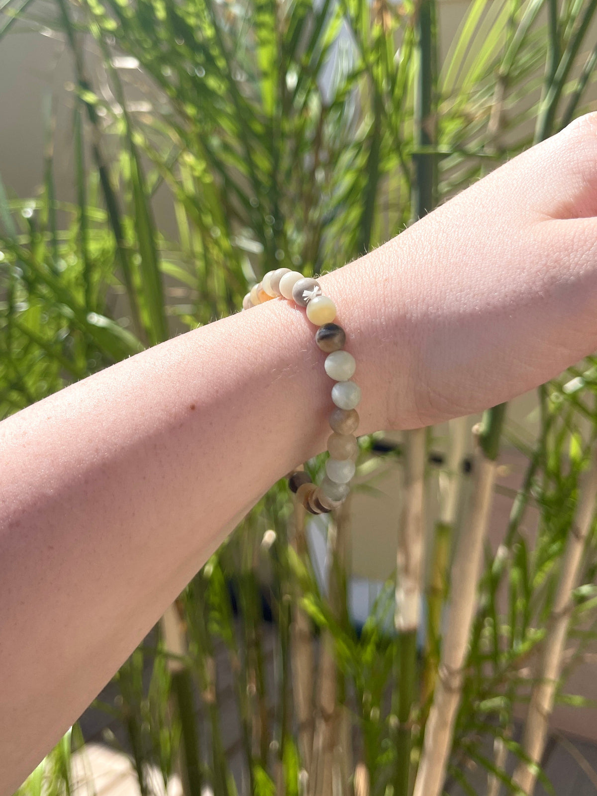 A hand wearing the Spellcrafter Sunstone Crystal Bracelet is extended in front of tall green plants on a sunny day.