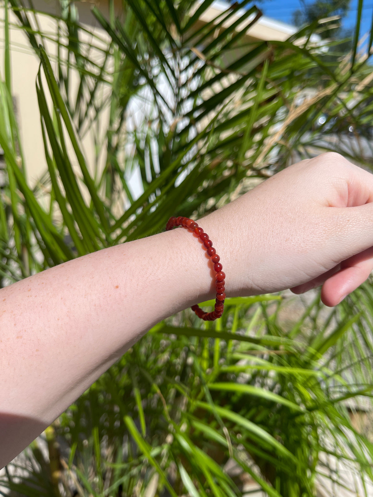A fair-skinned hand wears the Spellcrafter Carnelian Crystal Bracelet, held before green palm leaves with sunlight and shadows. A tan building and blue sky appear in the background.