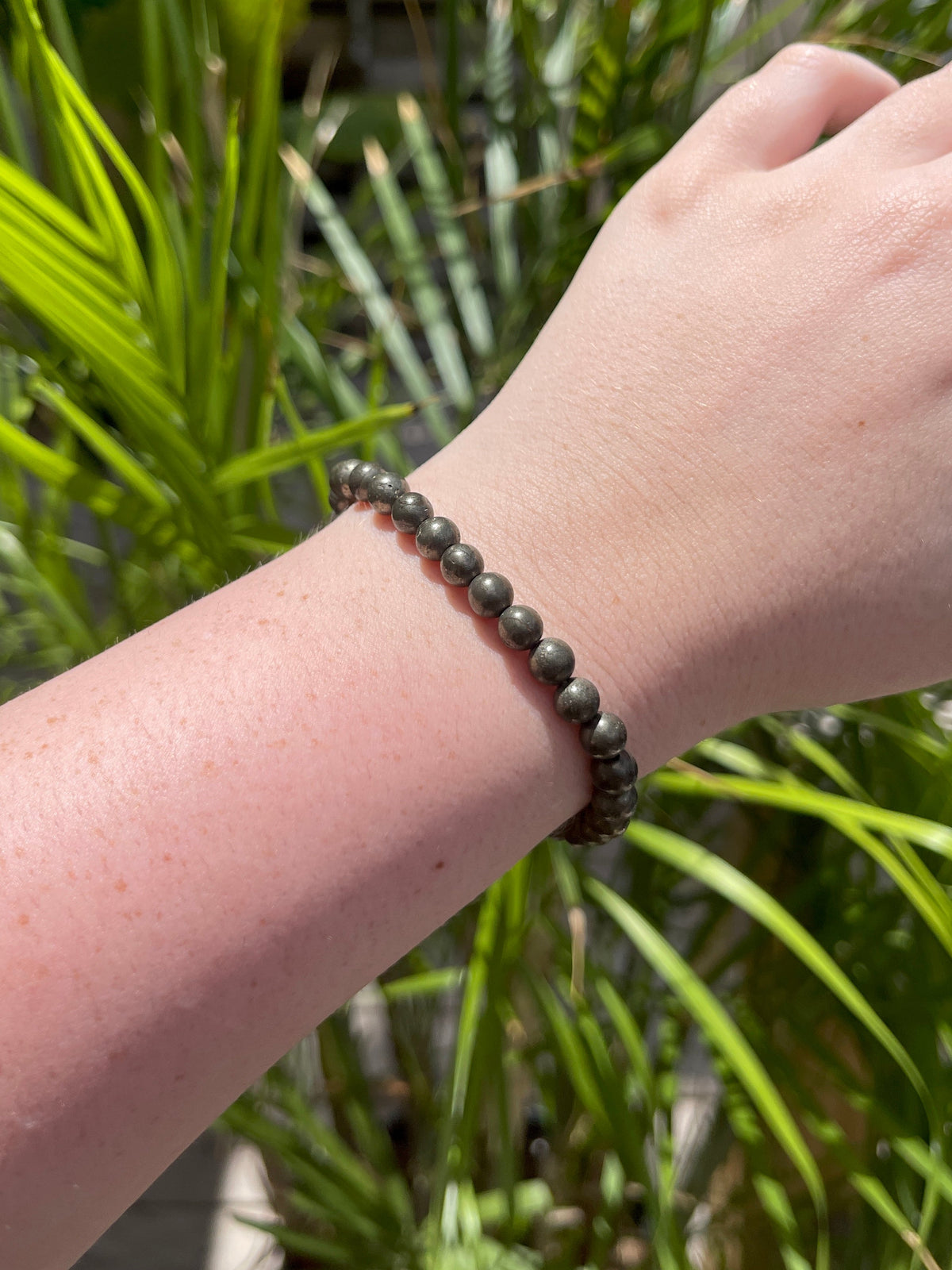 A fair-skinned hand wears the Spellcrafter Pyrite Crystal Bracelet with round, dark beads, shown in bright sunlight against tall green plants.