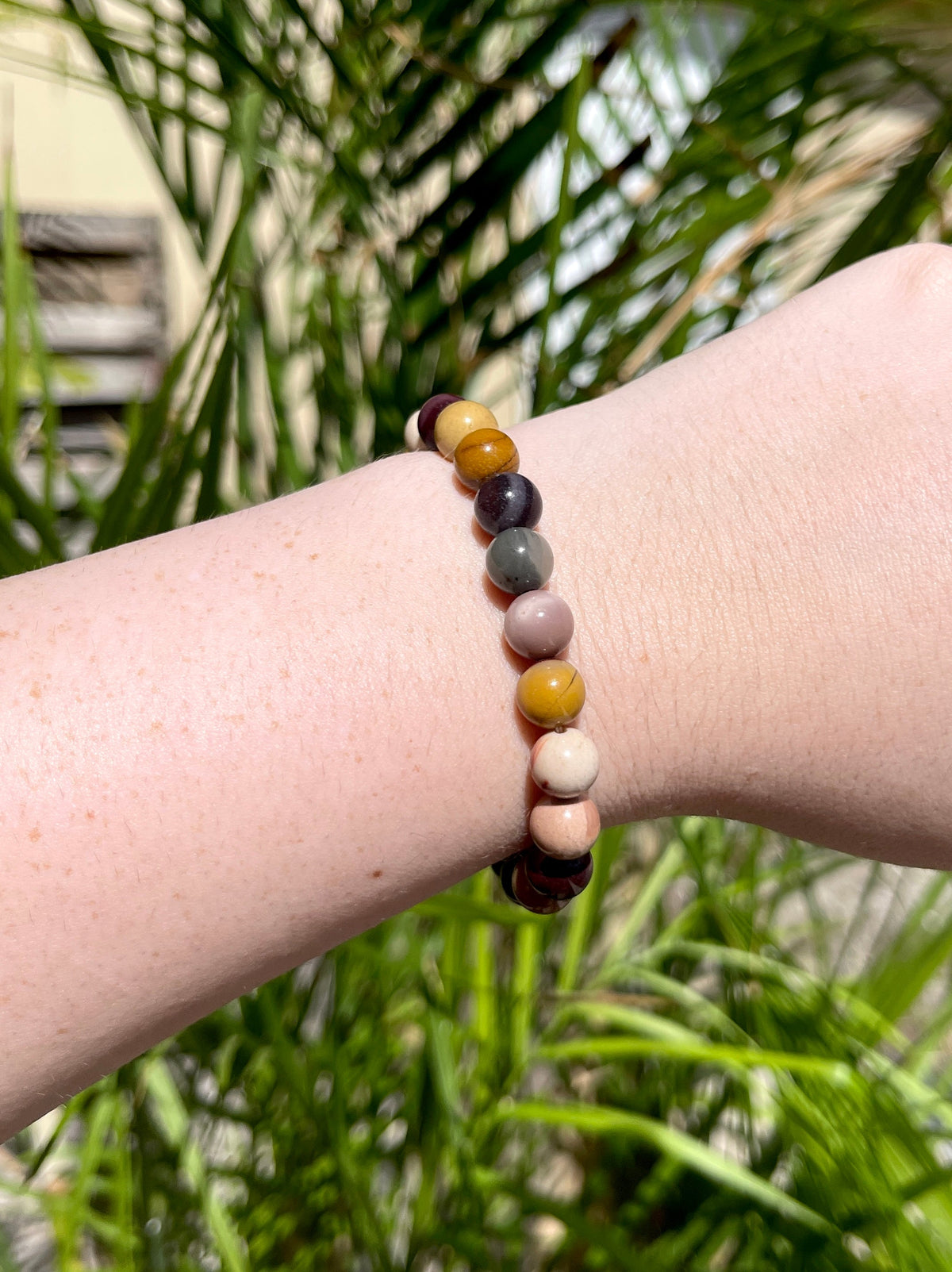A hand wears the Spellcrafter Mookaite Jasper Bracelet, featuring round, multicolored stones. Set outdoors with green leaves and sunlight in the background, the scene evokes healing energy and tranquility.