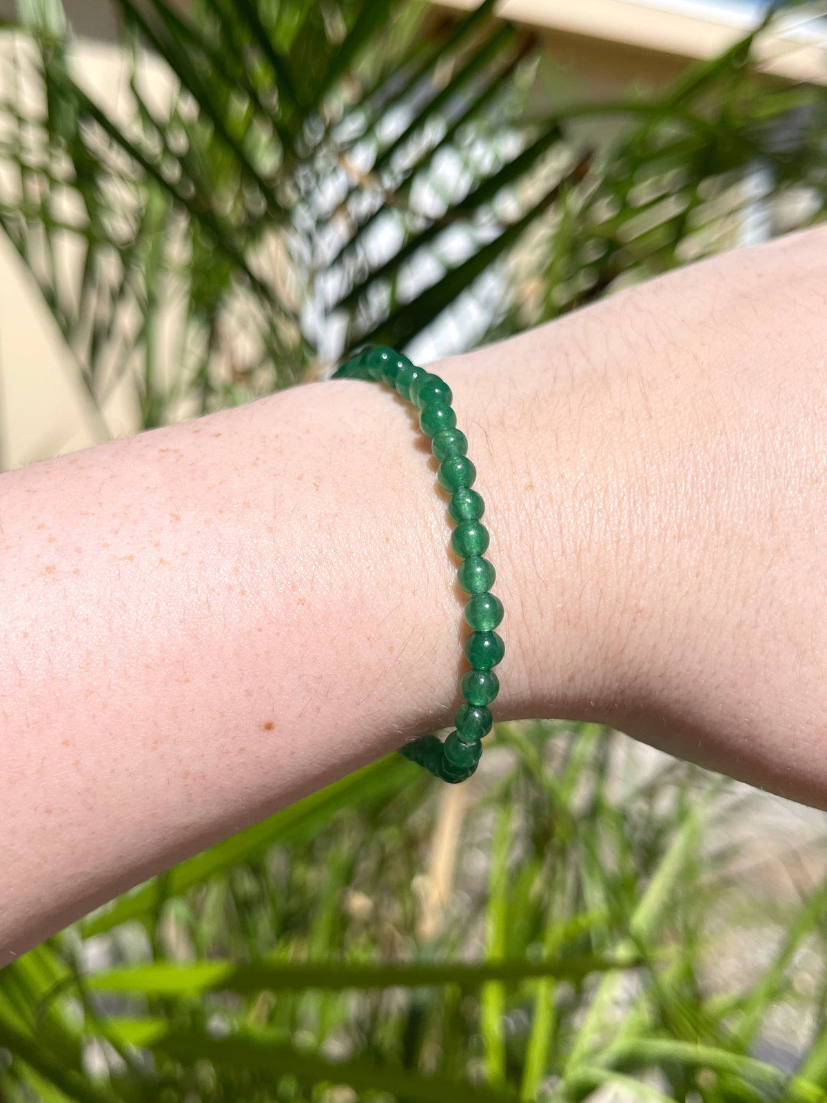 A close-up of a wrist wearing the Spellcrafter Green Aventurine Bracelet, with sunlight and palm leaves blurred in the background, radiating healing energy.