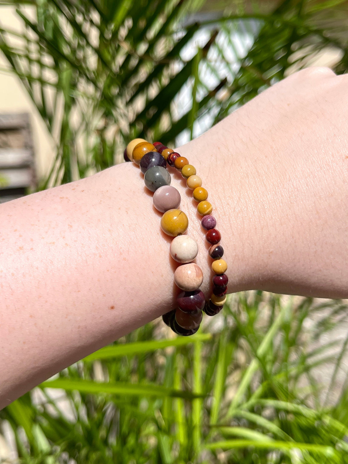 A hand wears two beaded bracelets—one is the Spellcrafter Mookaite Jasper Bracelet, featuring earthy yellow, brown, red, and gray tones—displayed outdoors among green plants and sunlight to highlight its natural healing energy.