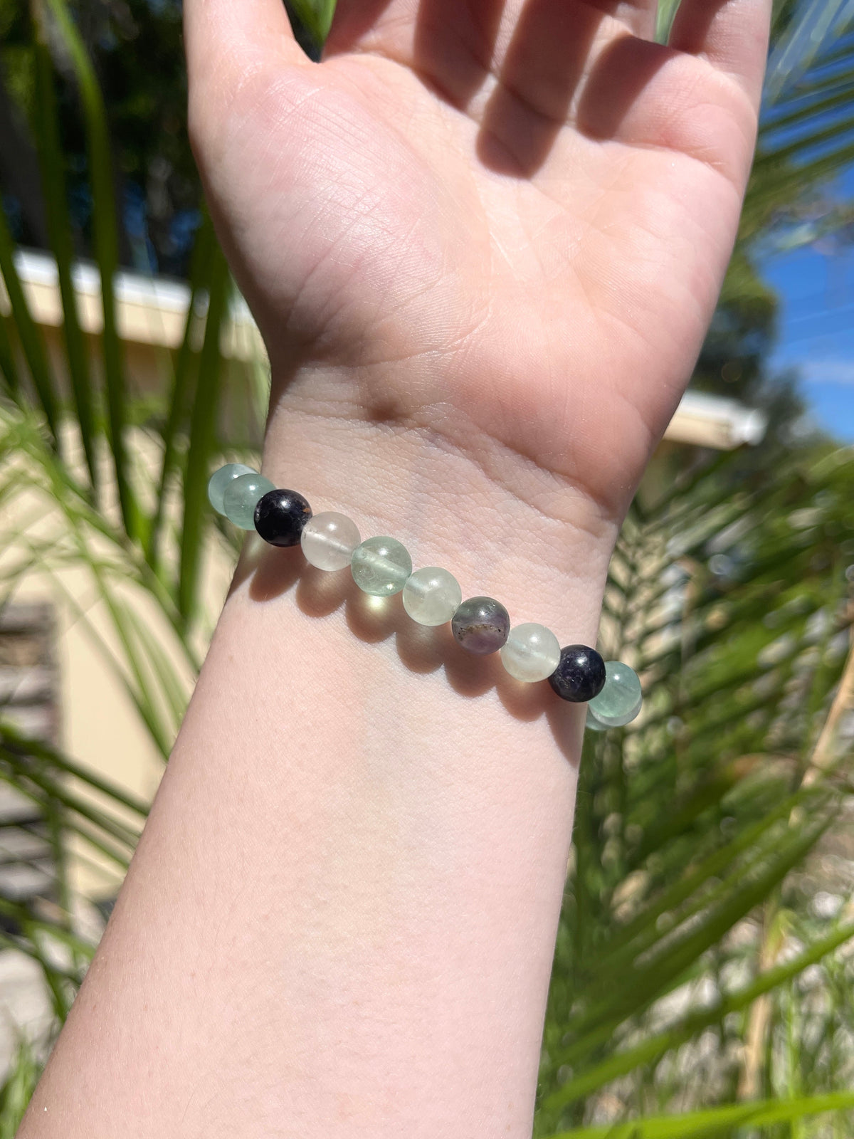 A hand wearing the Spellcrafter Fluorite Crystal Bracelet, featuring translucent light green beads and two black round beads, is held up outdoors with greenery and a building in the background.