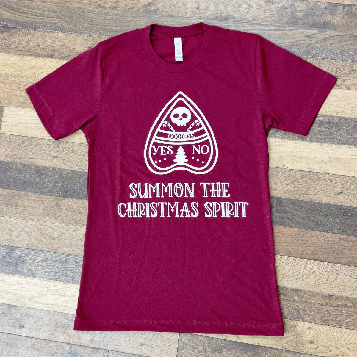 The Spellcrafter Summon The Spirit Christmas Tee is a maroon women's shirt with a white planchette design—YES, NO, GOOD BYE, skull, and Christmas tree—with "SUMMON THE CHRISTMAS SPIRIT" below on soft Heather CVC fabric.