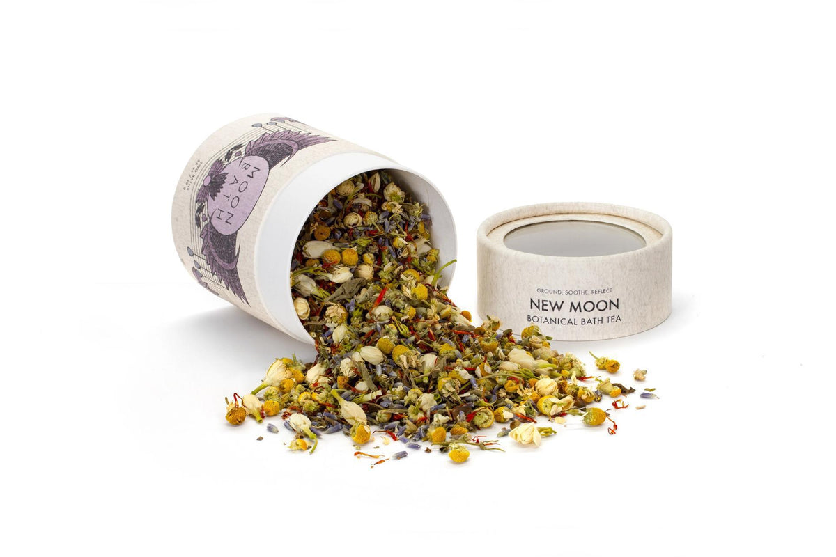 A Spellcrafter New Moon Bath Tea cylinder lies on its side with the lid off, spilling a vibrant blend of dried flowers and herbs—like lavender and chamomile—across a white surface.