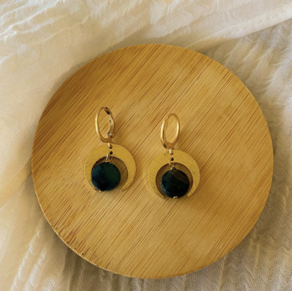 The Spellcrafter Apatite Quarter Moon Earrings feature gold hoops with textured circular pendants and dark green gemstone accents, inspired by moon phase jewelry, shown on a round wooden plate with a white fabric background.