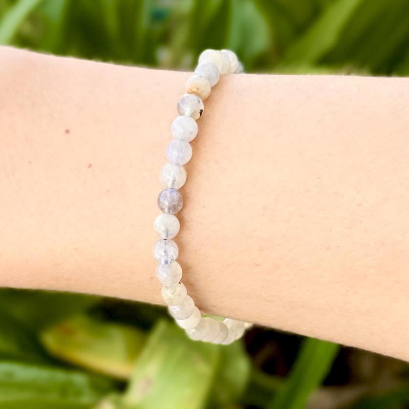 A close-up of a wrist wearing the Spellcrafter Labradorite Crystal Bracelet, featuring small, round, light stones, set against a blurred green background.