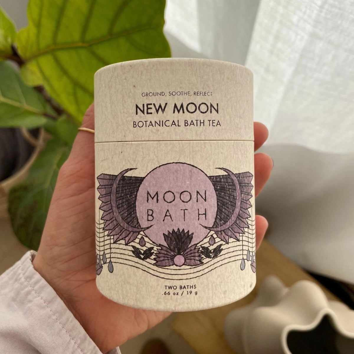 A hand holds Spellcrafter’s New Moon Bath Tea—an elegant cylinder with purple and black celestial designs, perfect for a soothing soak with calming lavender and chamomile. A plant appears in the background.