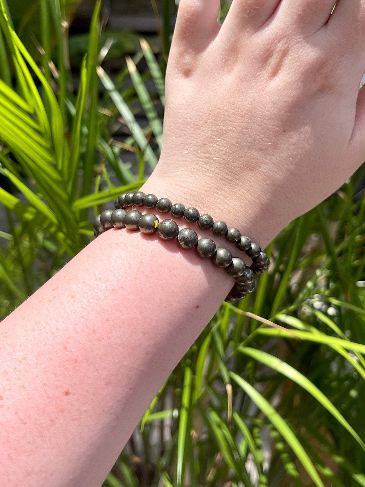 A hand wears two dark Spellcrafter Pyrite Crystal Bracelets outdoors, with green leafy plants softly blurred in the background.