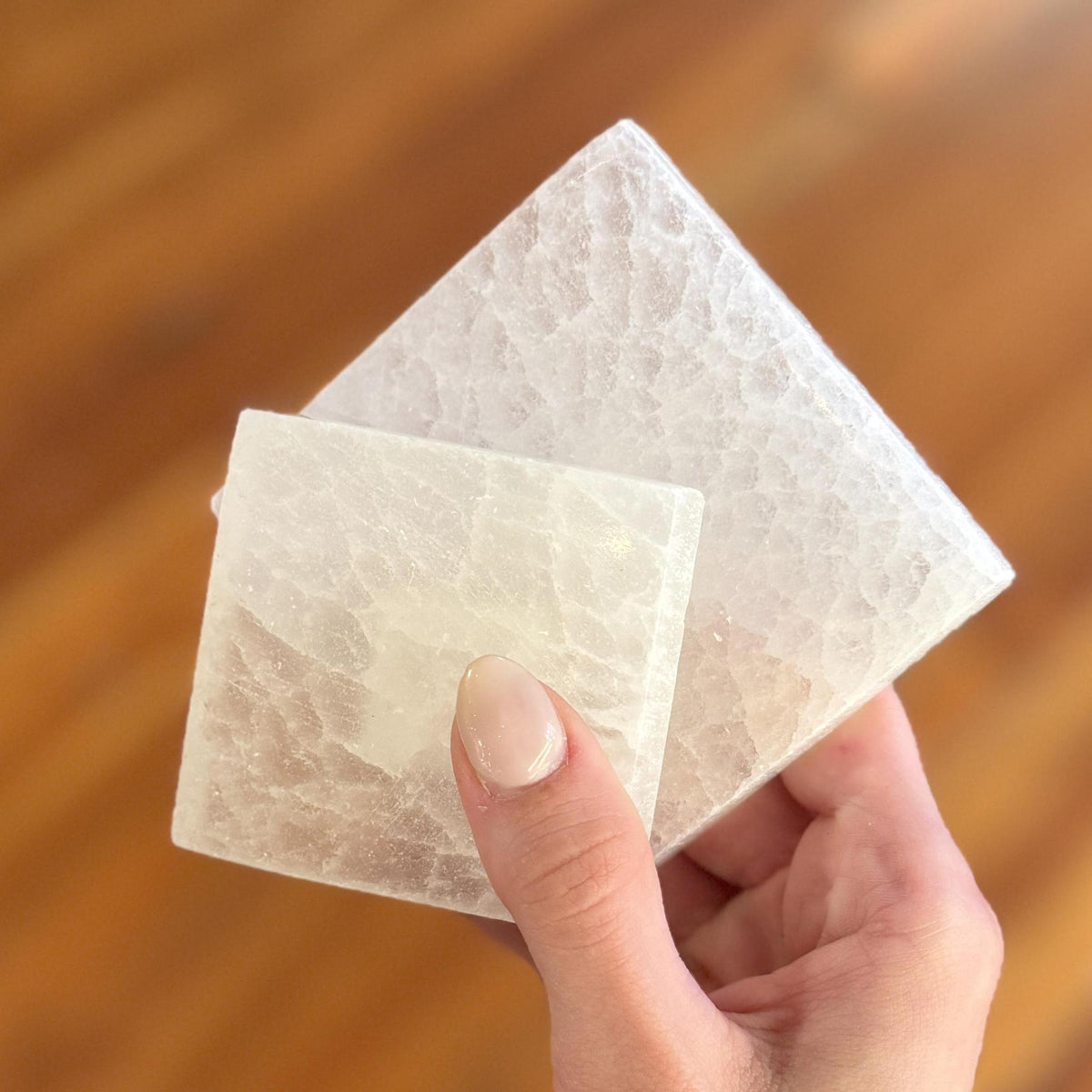 A hand displays two Spellcrafter Selenite Square Plates of varying sizes, their translucent surfaces perfectly balanced against a softly blurred wooden background.