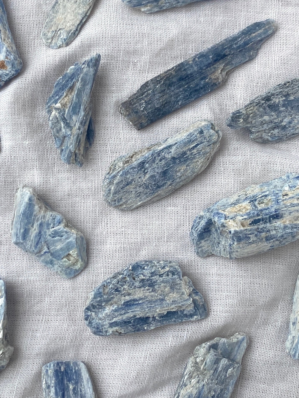 Spellcrafter Rough Kyanite Chunks are scattered on a white fabric background, showcasing their natural texture and blue-gray shades—ideal for meditation and relaxation.