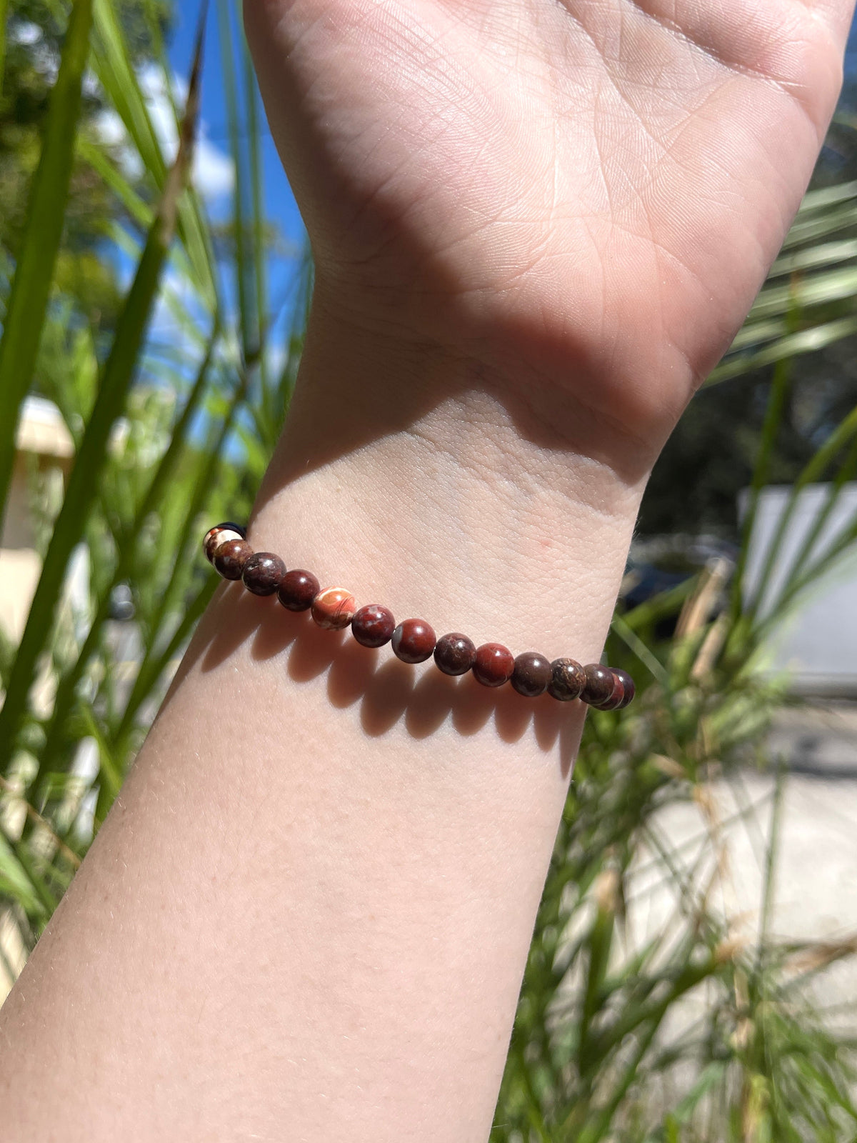 A hand with the palm facing up wears the Spellcrafter Jasper Bracelet, made of round, reddish-brown beads. Green plants and sunlight create a vibrant outdoor background.