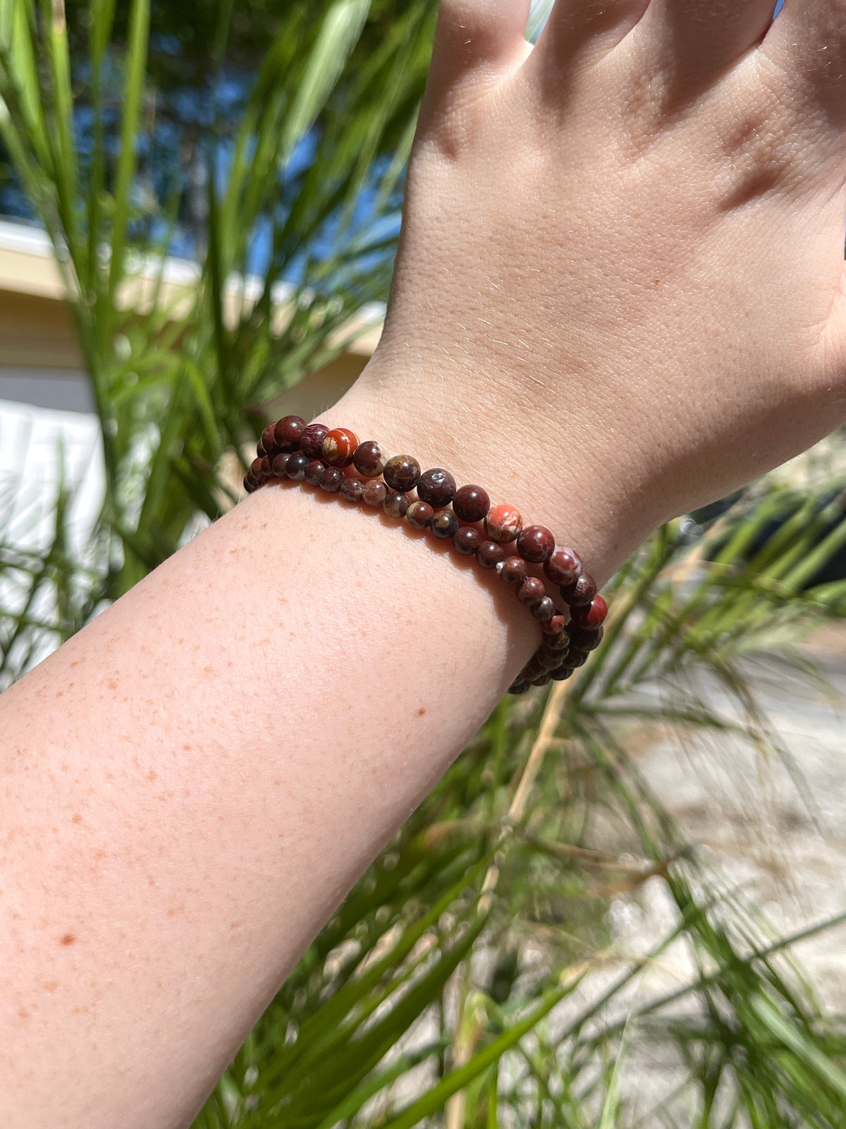 A fair-skinned hand and forearm wear two Spellcrafter Jasper Bracelets, shown outdoors among green plants and sunlight.
