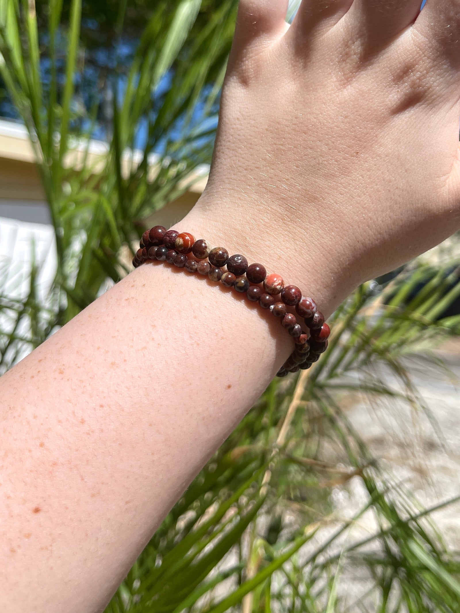 A fair-skinned hand and forearm wear two Spellcrafter Jasper Bracelets, shown outdoors among green...