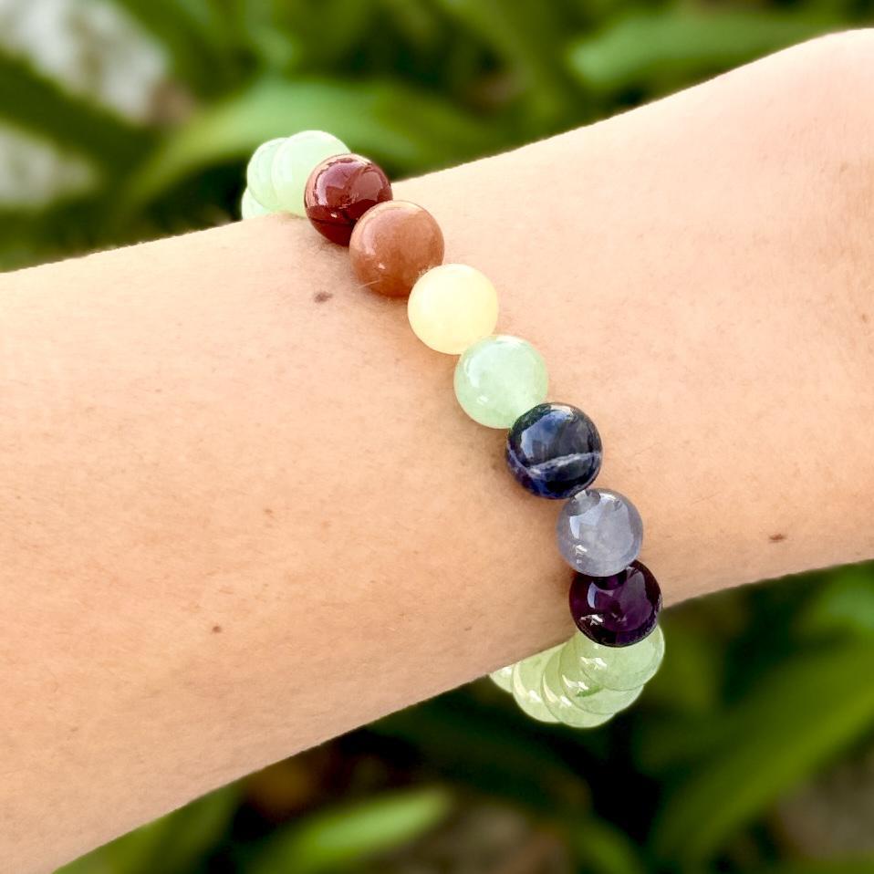 A close-up of a wrist wearing the Spellcrafter Chakra Stone Bracelet, featuring round, polished stones in green, yellow, red, blue, and purple hues against a blurred green background.