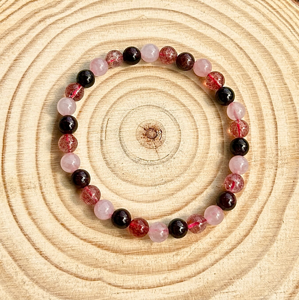 The Spellcrafter Heart Chakra Trio Crystal Bracelet features alternating pink, dark red, and crackled red beads, arranged in a circle on a wooden surface with tree rings—ideal for heart chakra support.