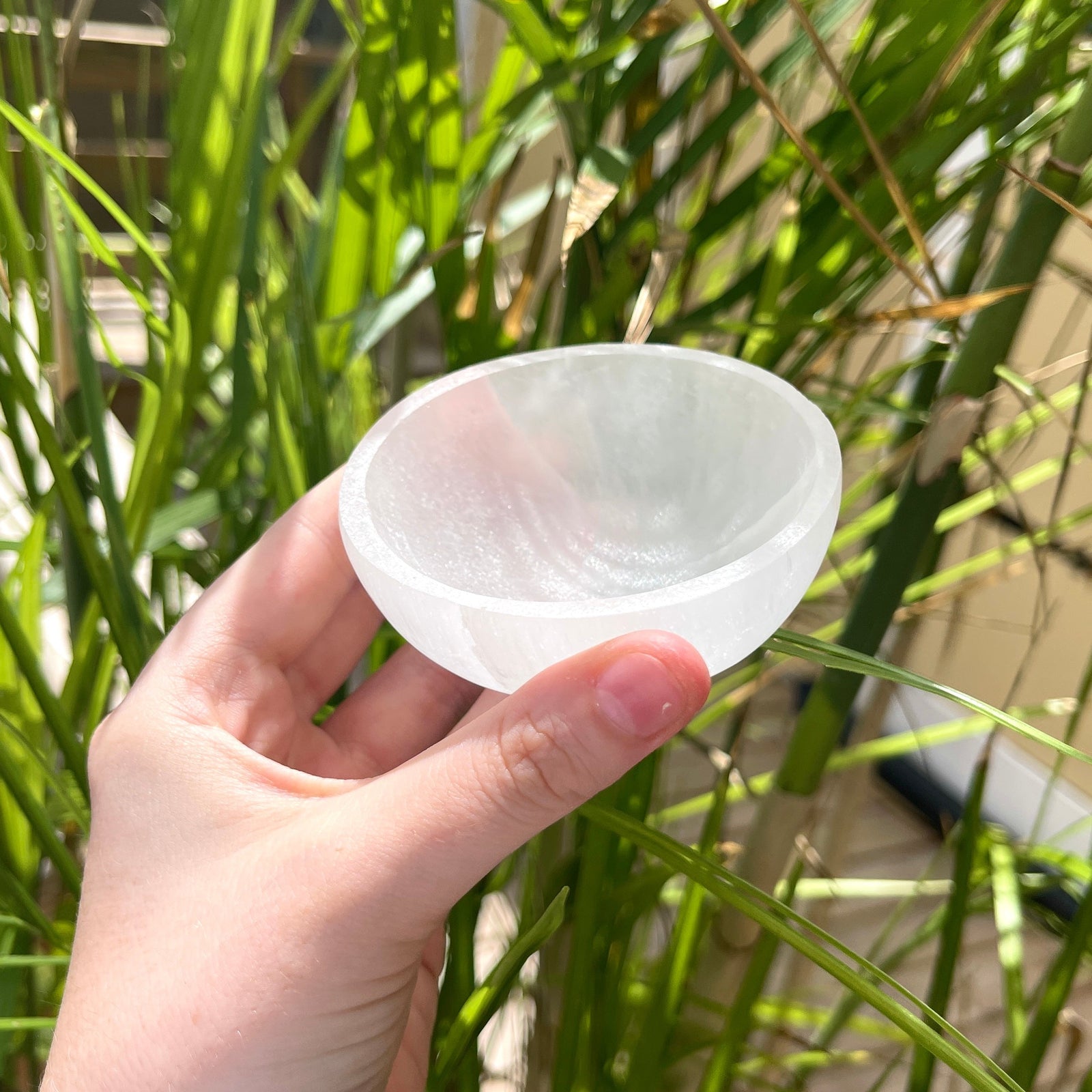 A hand displays the Spellcrafter Small Selenite Bowl outdoors against tall green grass—ideal for...