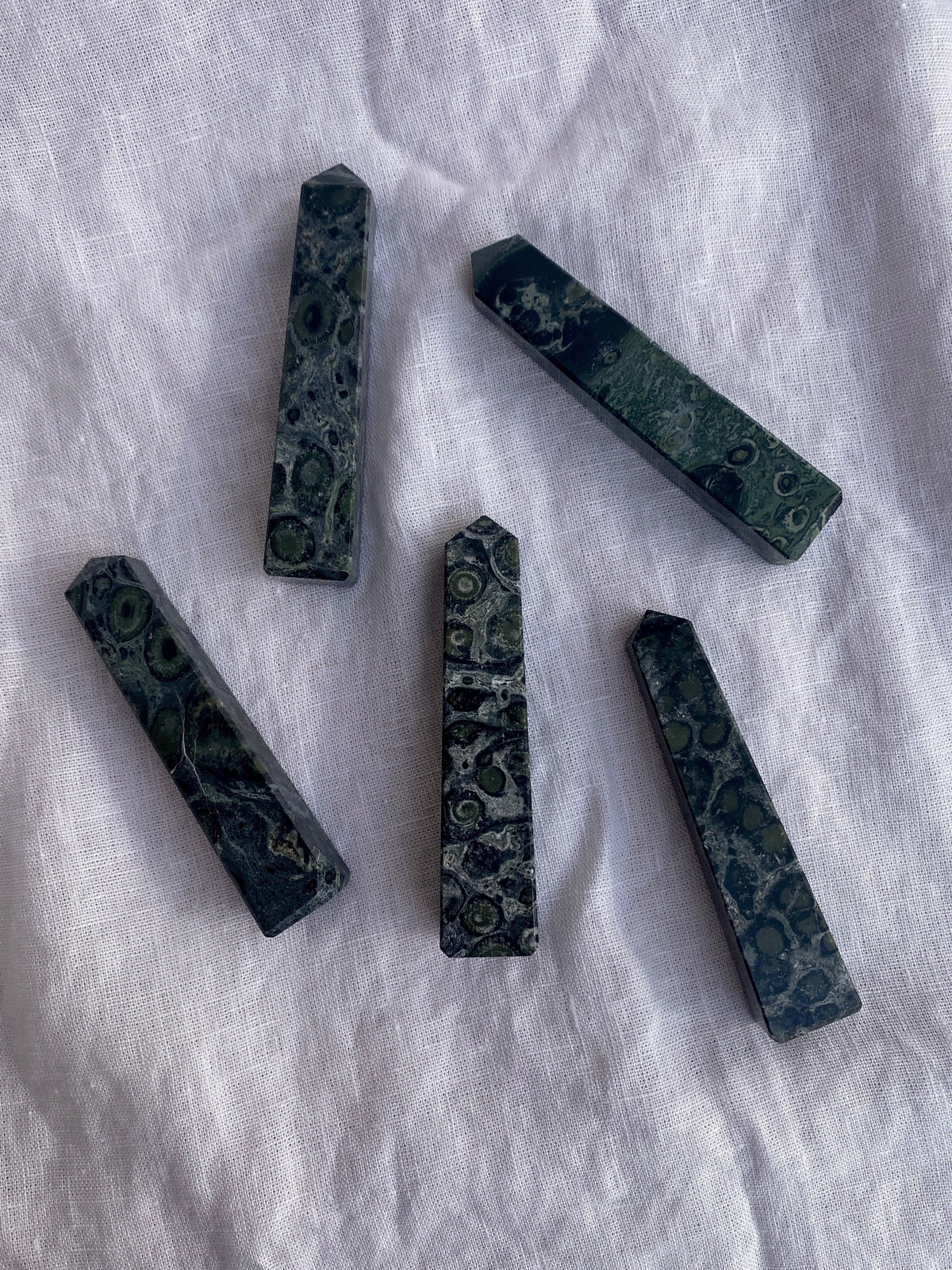 Five Spellcrafter Large Kambaba Jasper Crystal Points with rounded, circular patterns are arranged on a white, textured fabric background.