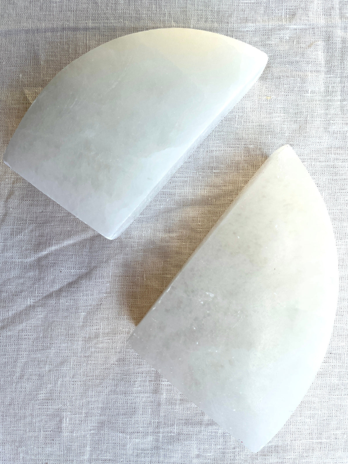 Spellcrafter Selenite Bookends—two smooth, white, semi-circular pieces with opposite curved edges—rest on a light textured fabric surface, adding a refined, polished touch to any home décor.