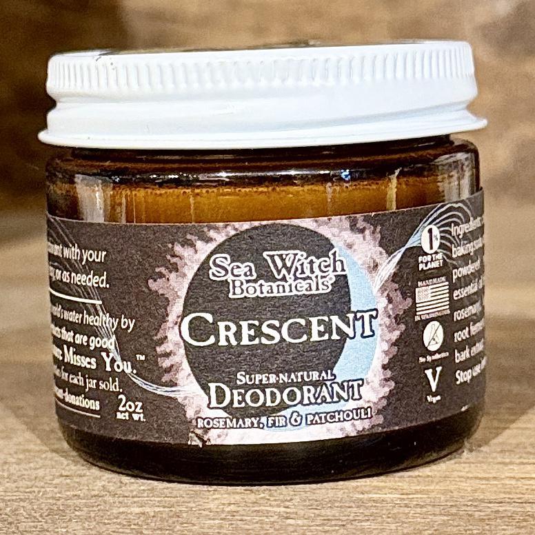A brown glass jar of Spellcrafter SuperNatural Deodorant with a white lid, featuring rosemary, fir, and patchouli scents, rests on a wooden surface.
